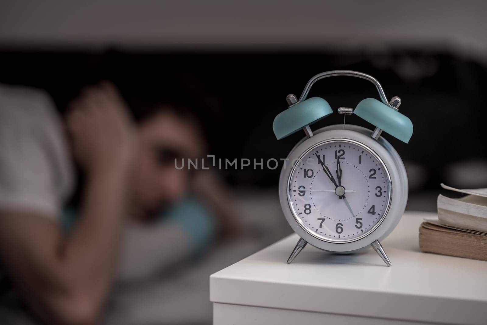 White alarm clock on the bedside table. Young tries to sleep in the background, insomnia