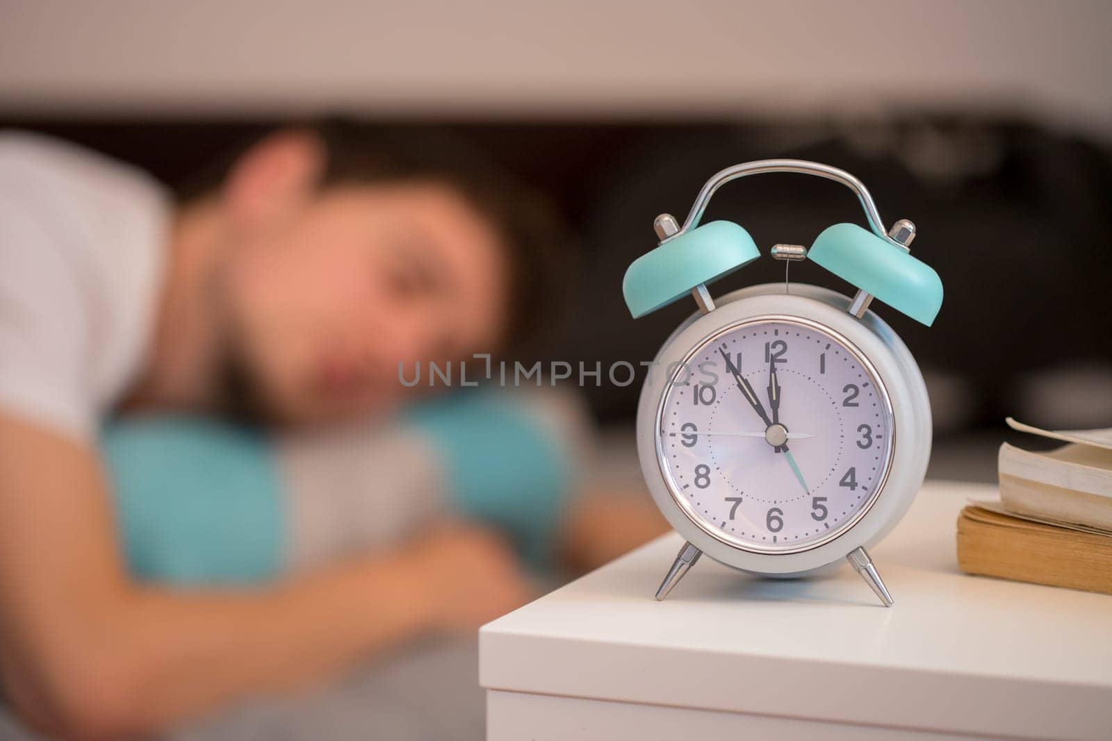White alarm clock on the bedside table. Young man sleeps in the background.