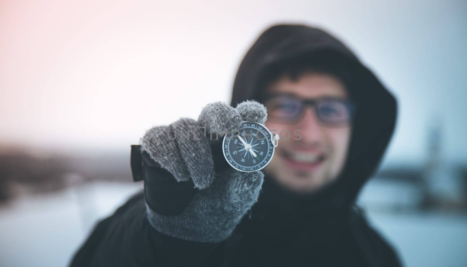 Young smiling man holds up a compass. Wintertime, gloves and hood.