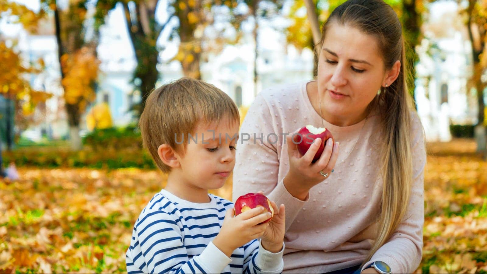Portrait of little todler boy with mother eating red apples in autumn park.
