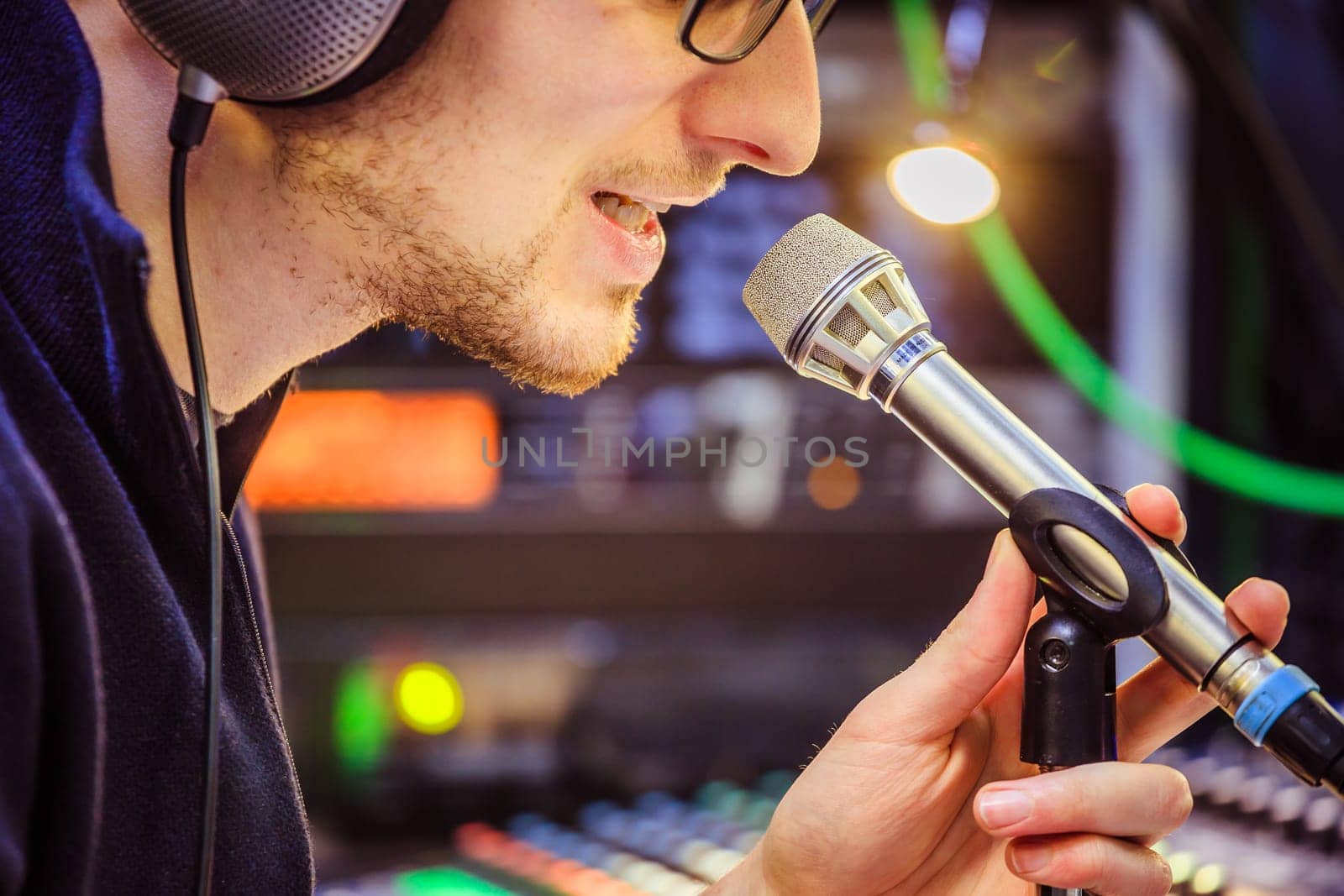 Young man is sitting in the recording studio and talks into the microphone, buttons and equipment in the blurry background