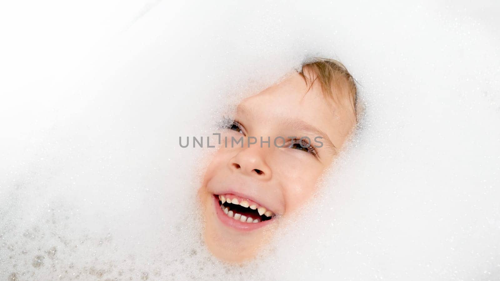 Portrait of happy laughing boy covered in soap foam swimming and playing in bath.