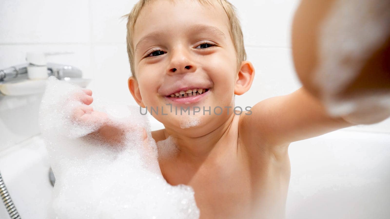 Portrait of cute toddler boy covered in soap foam taking bath and looking in camera.