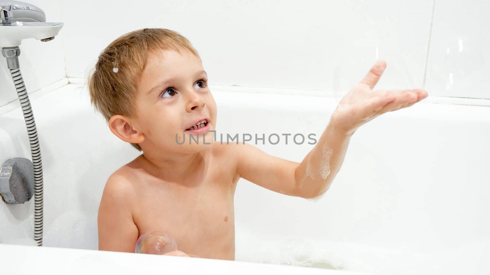 Portrait of cute boy catching soap bubble while washing in bath.