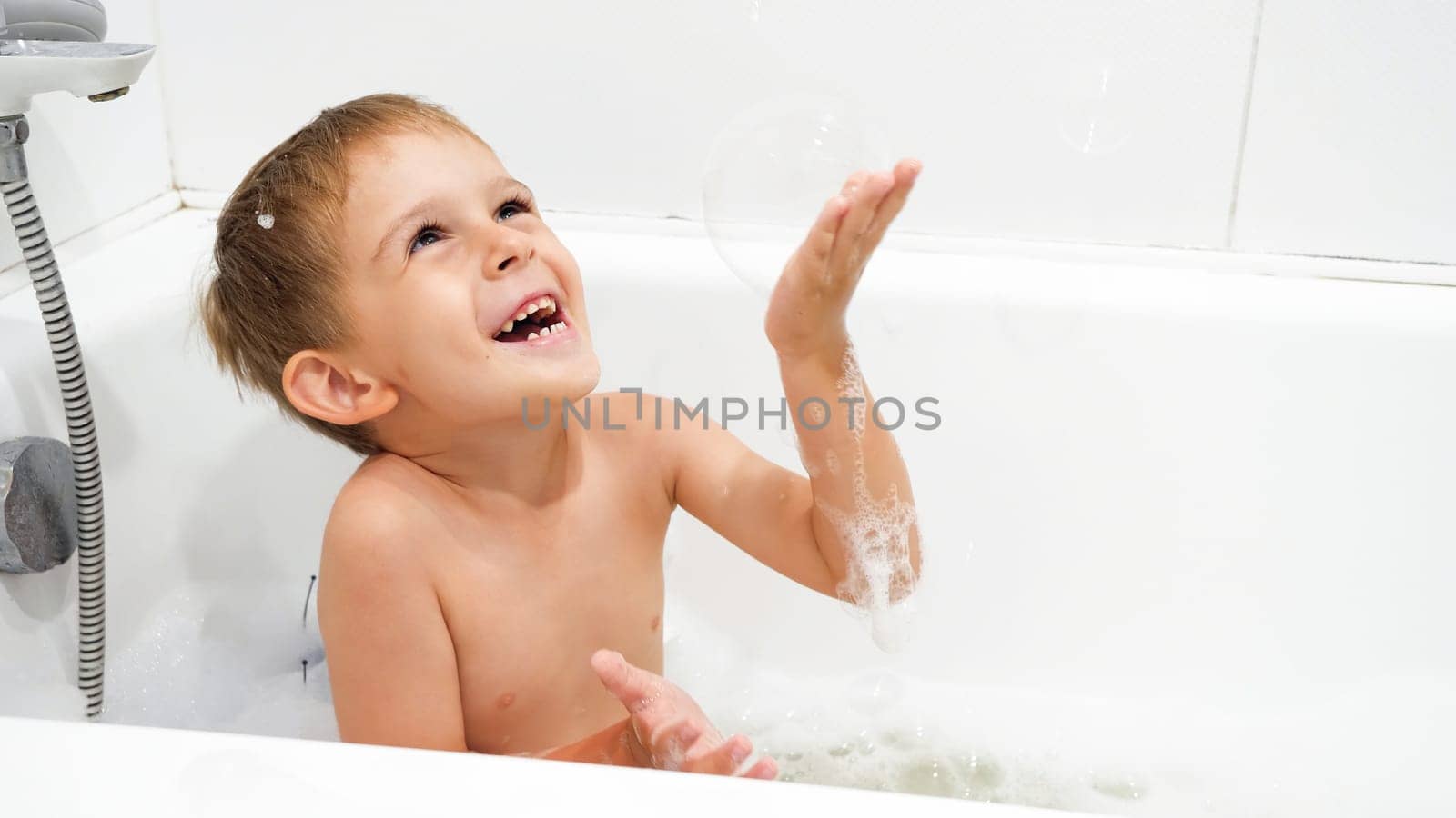 Happy smiling boy playing with soap bubbles and foam in bath.