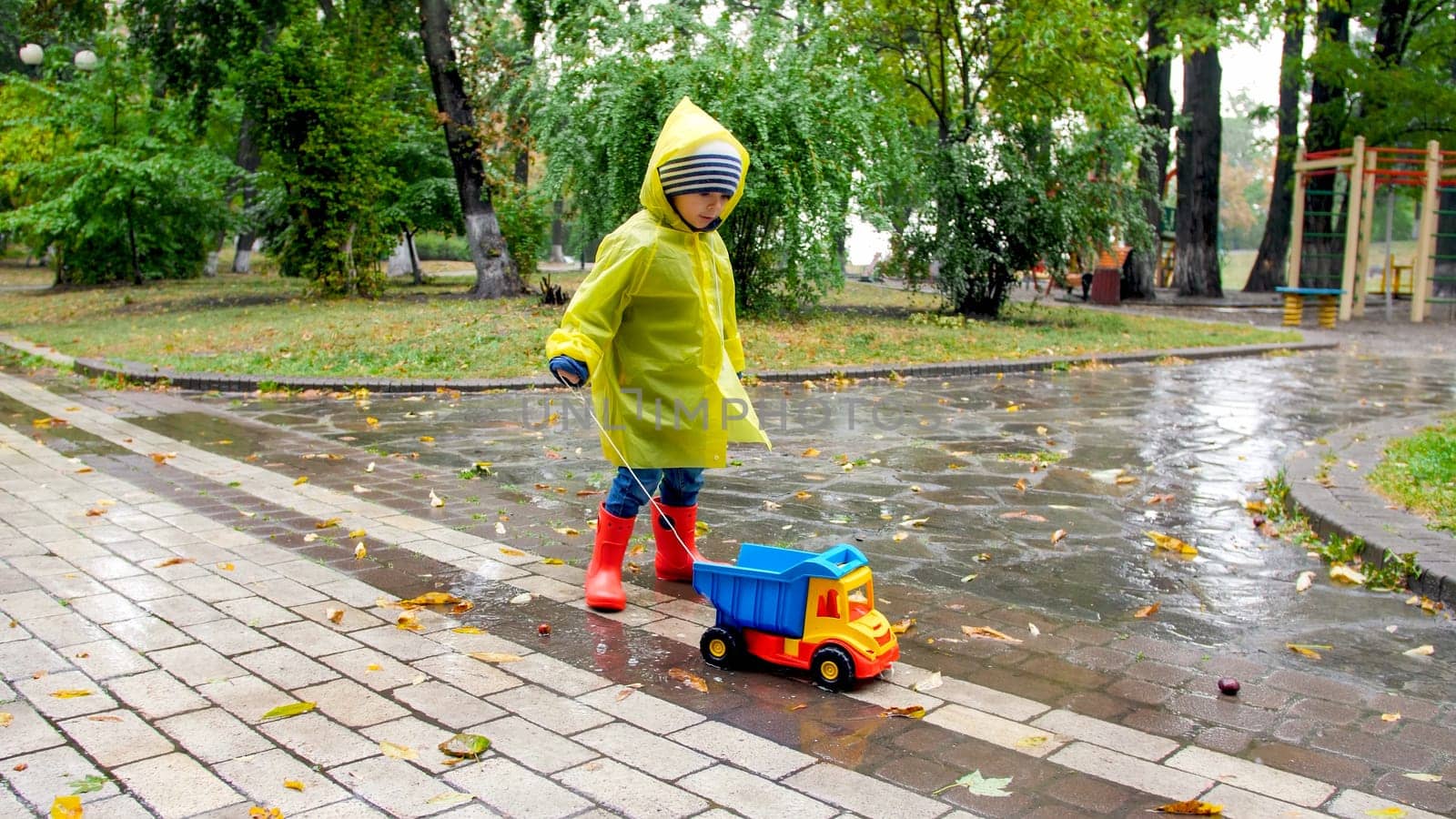Cute little boy in raincoat and rubber boots blaying with toy truck in big puddle at park.