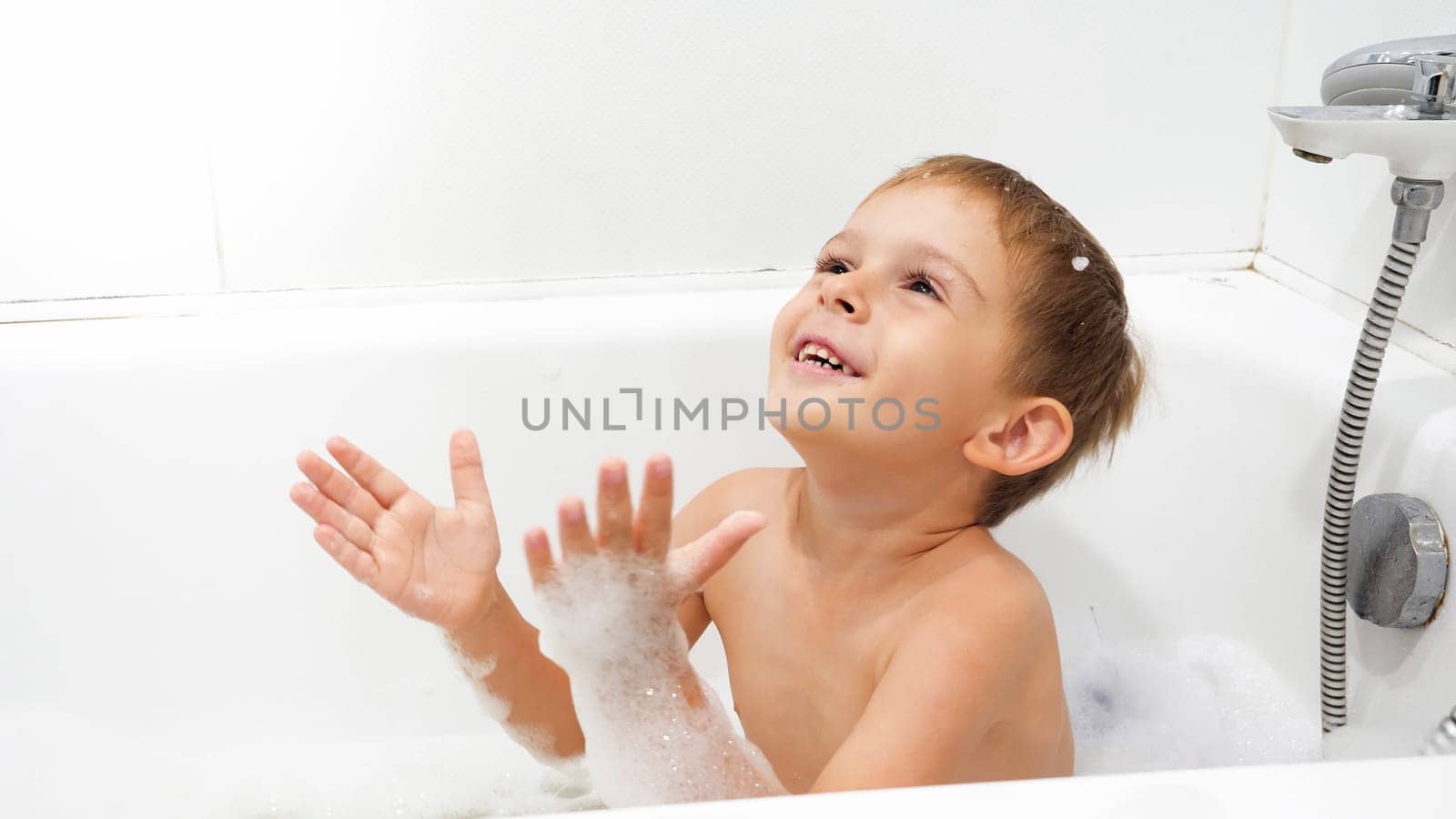 Portrait of cheerful smiling little boy enjoying washing in bath with soap foam.
