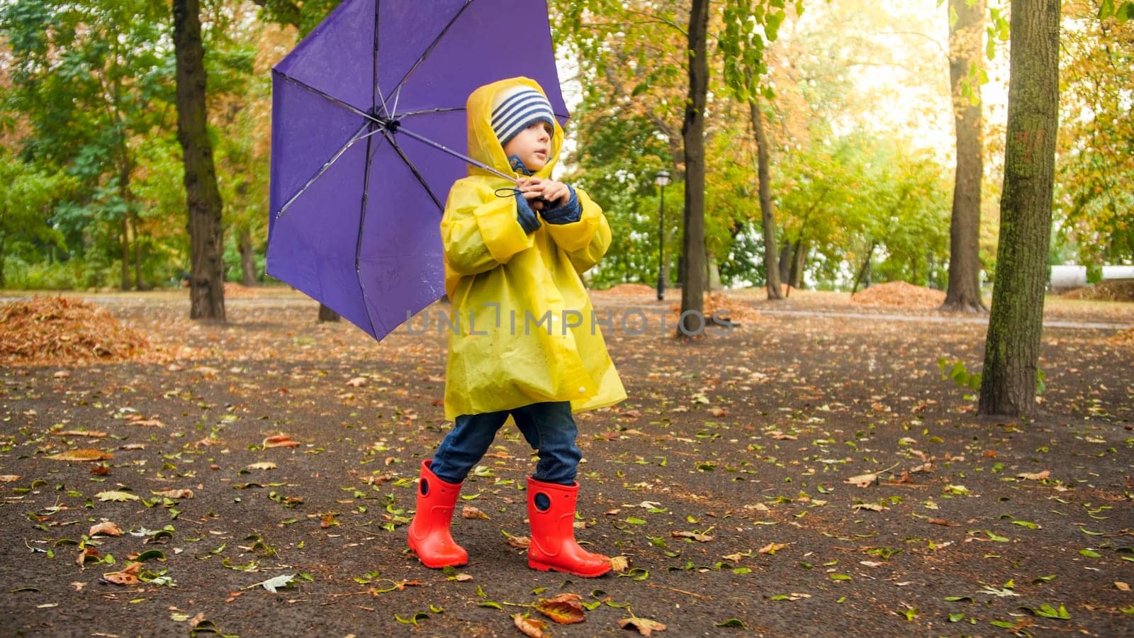 Cute child in boots and raincoat walking with umbrella at autumn park.