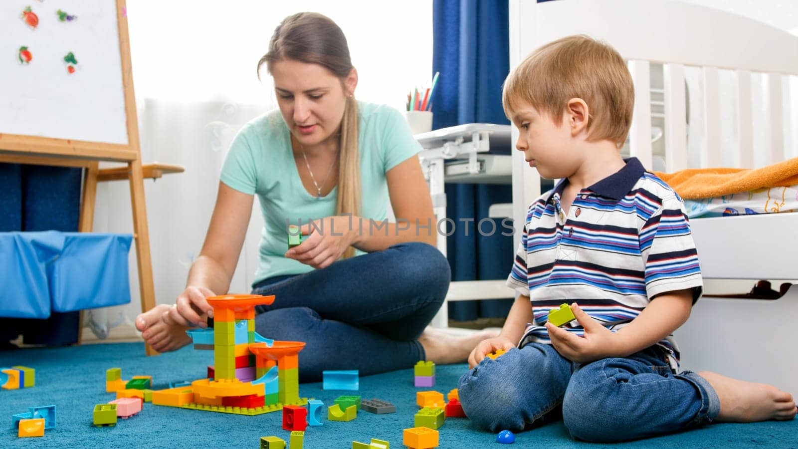 Young mother helping her little son assembling toy constructor on carpet at playroom.
