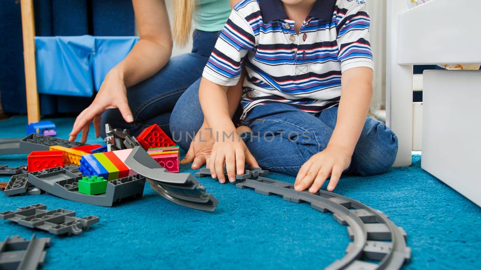 Closeup image of little toddler boy wth young mother assembling toy railroad on floor.