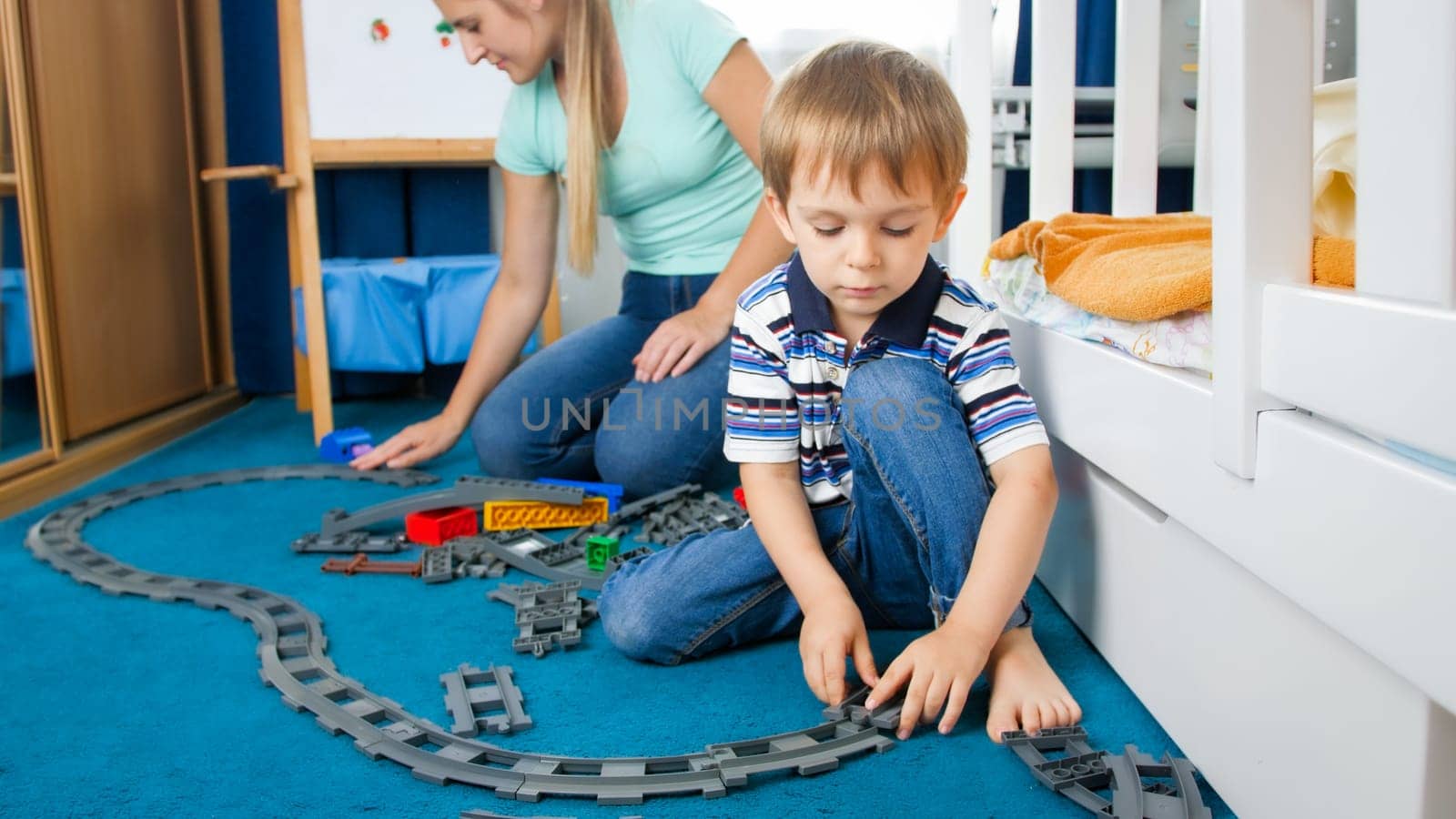Cute little boy and his young mother playing with toy railroad on floor at bedroom.