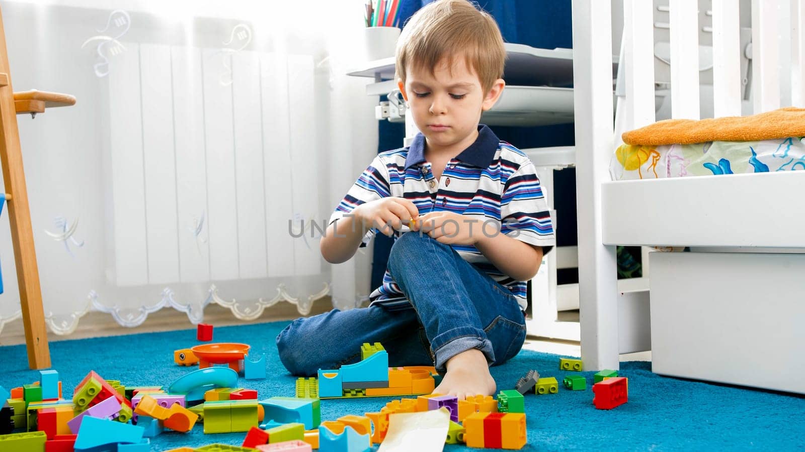 Portrait of little concentrated boy palying on carpet with toy blocks.