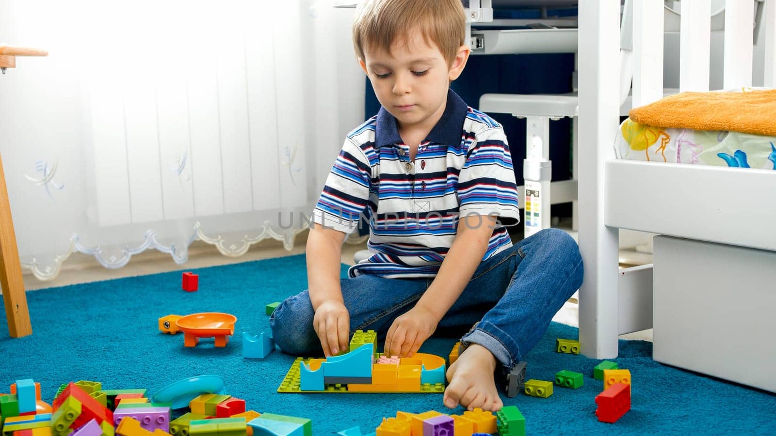 Portrait of little toddler boy sitting on floor in bedroom and playing with toy constructor.