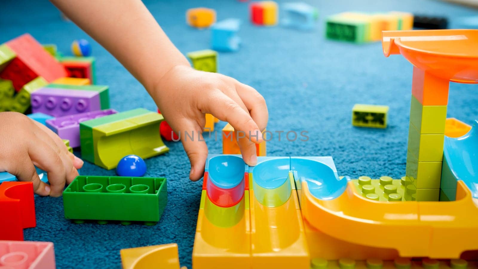 Closeup image of child's hand building toy track with colorful constructor blocks on floor.