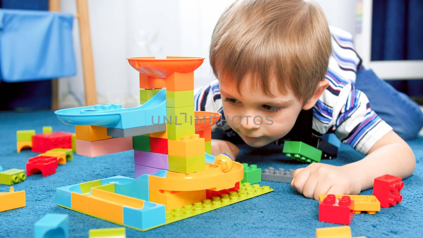 Portrait of little concentrated boy lying on floor and looking on toy marble run game.