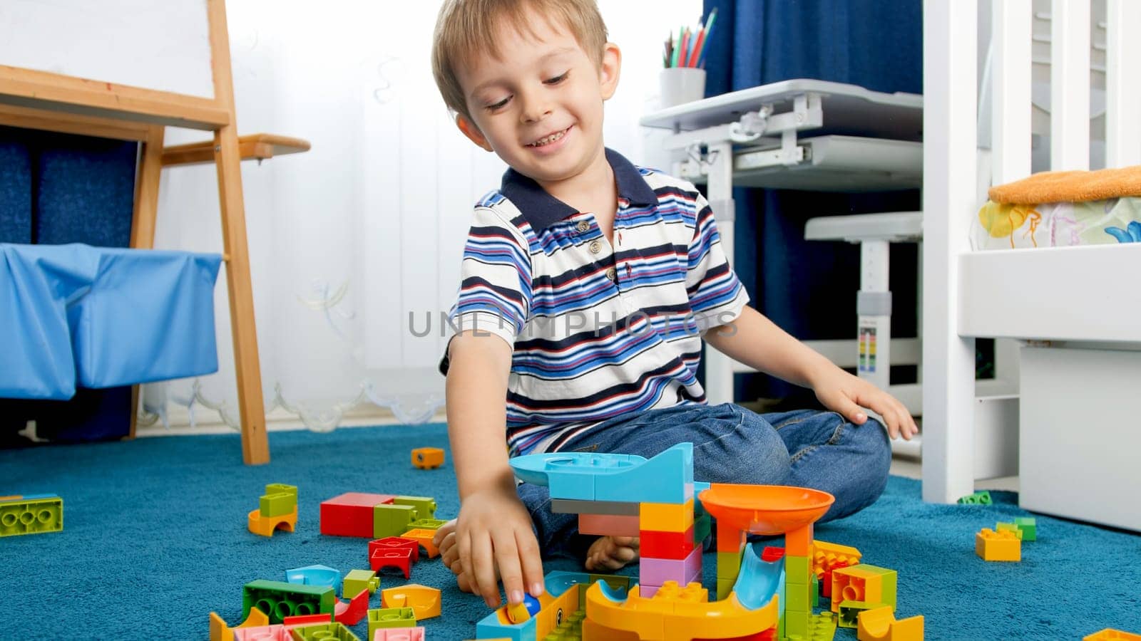 Portrait of happy smiling boy playing with colorful constructor blocks on carpet in playroom.