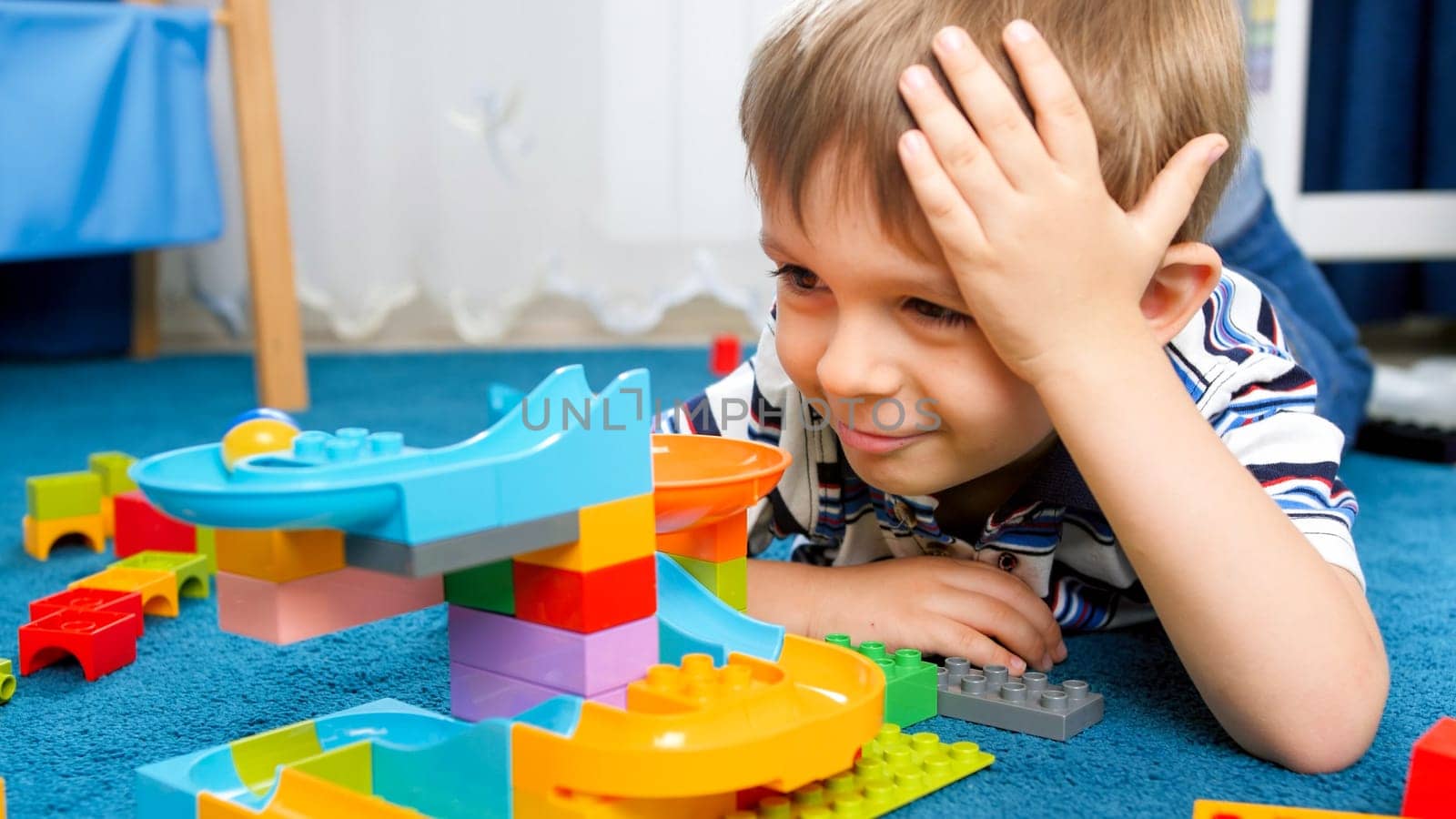 Portrait of happy smiling little boy lying on carpet at his room and looking on colorful toy constructor.