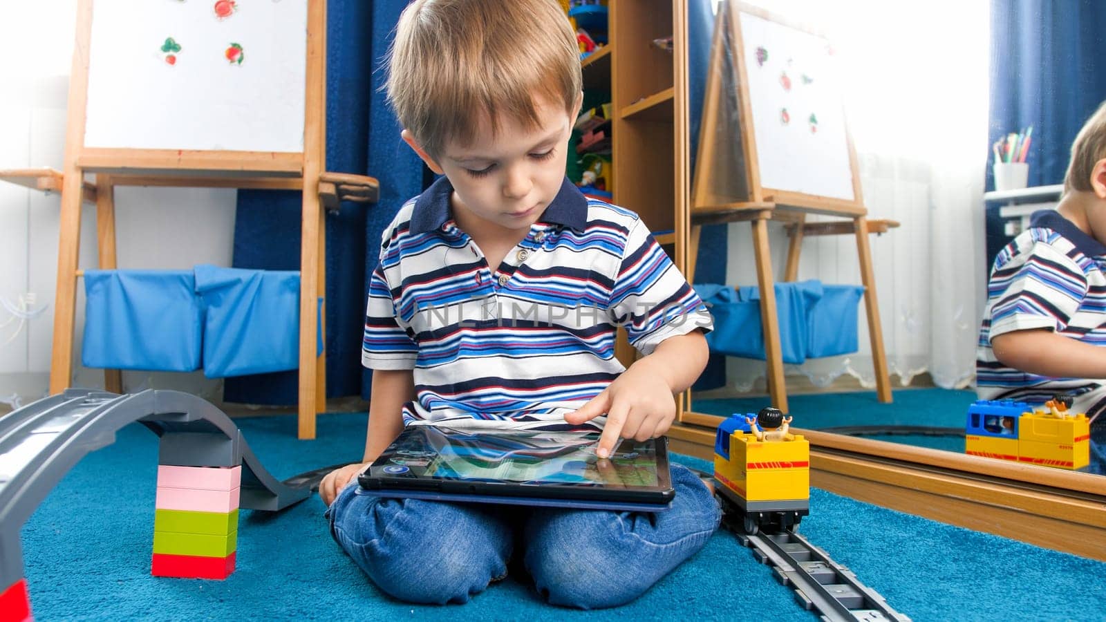 Little boy sitting in messy room and playing games on tablet computer.