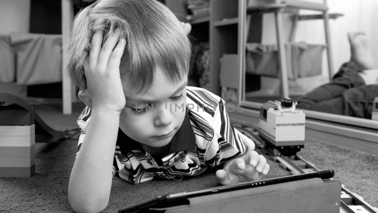 Black and white portrait of little boy lying on floor at living room and using tablet computer.