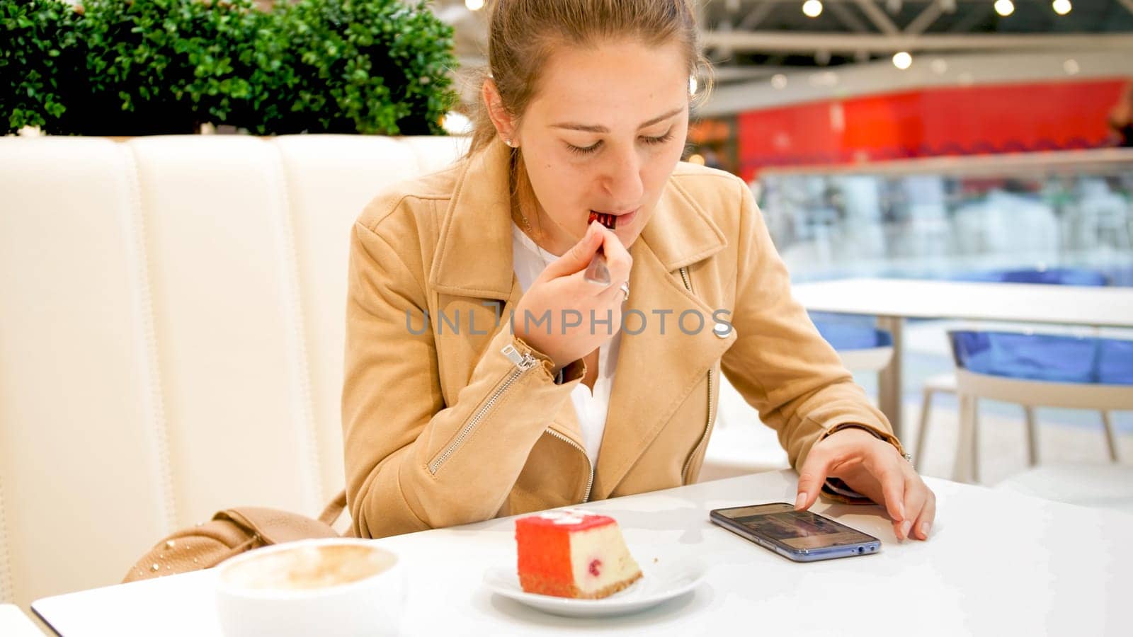 Portrait of beautiful young woman eating dessert and reading news on smartphone at cafe.