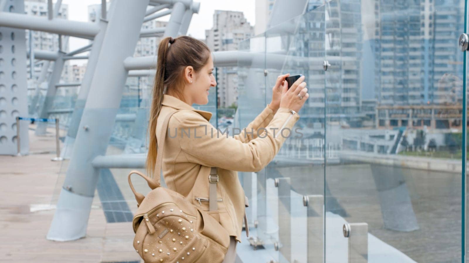 Portrait of beautiful young woman making photograph of river and city skyline on smartphone from glass bridge.