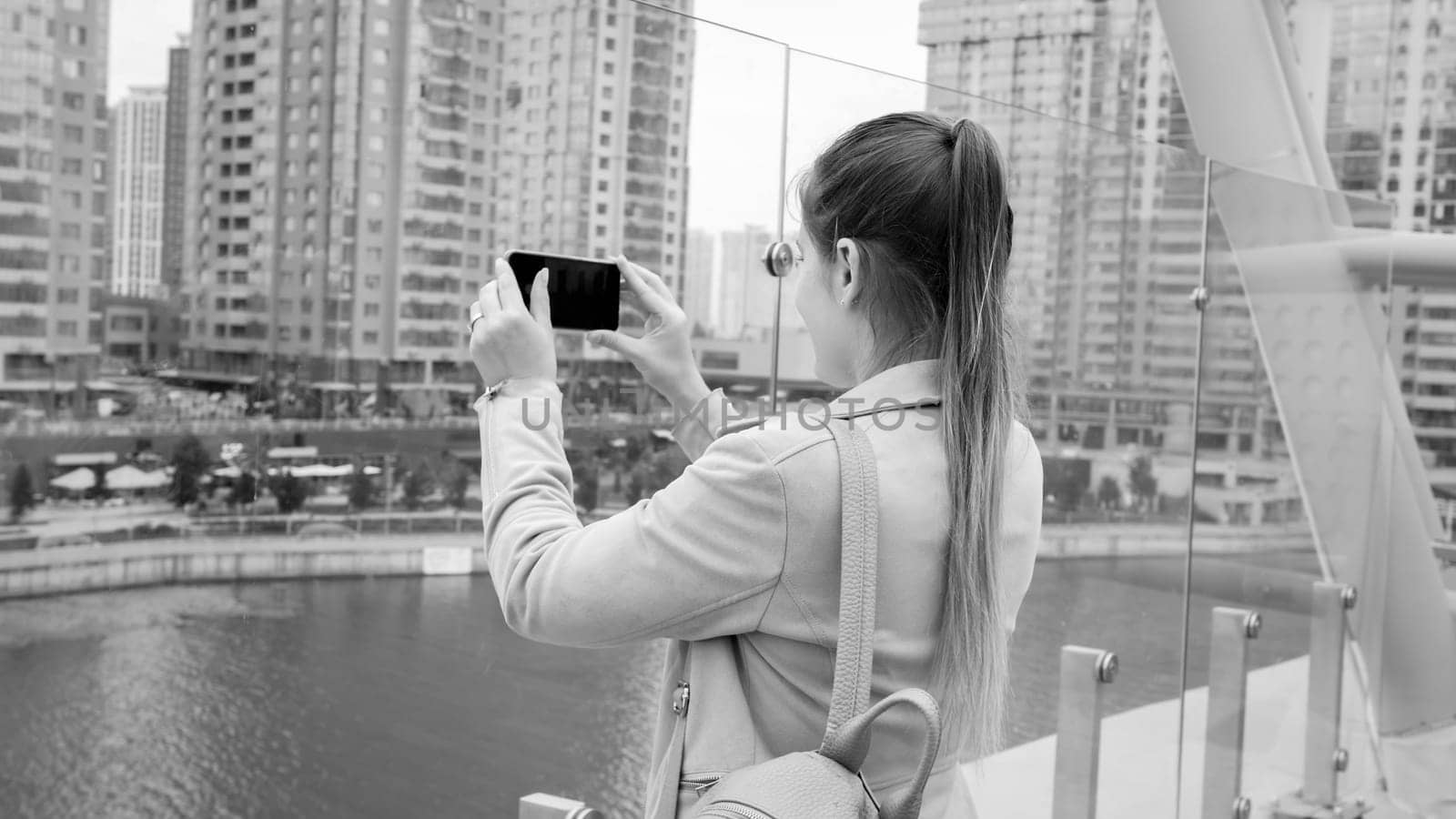 Black and white photo of young woman taking images of city skyline and river on smartphone.