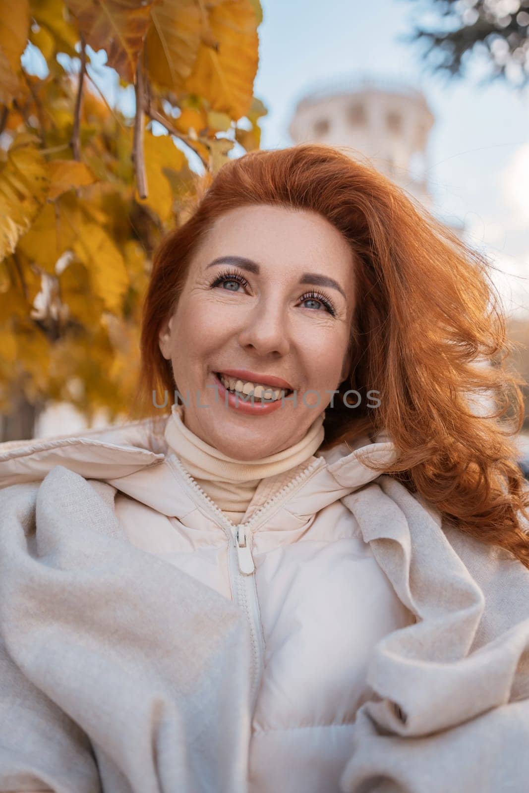 Smiling redhead woman in a beige coat, windswept hair, autumn leaves in the background. Joyful autumnal portrait.