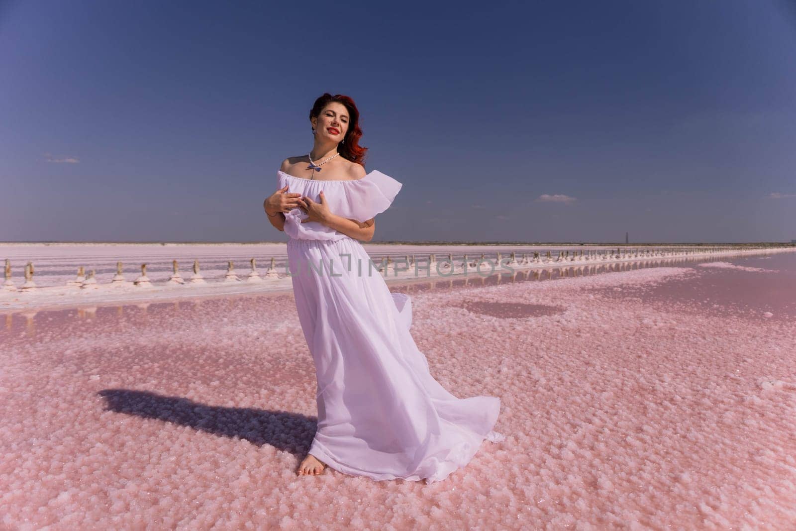A woman in a white dress stands on a pink salt flat, her hair flowing in the wind, with a clear blue sky above.