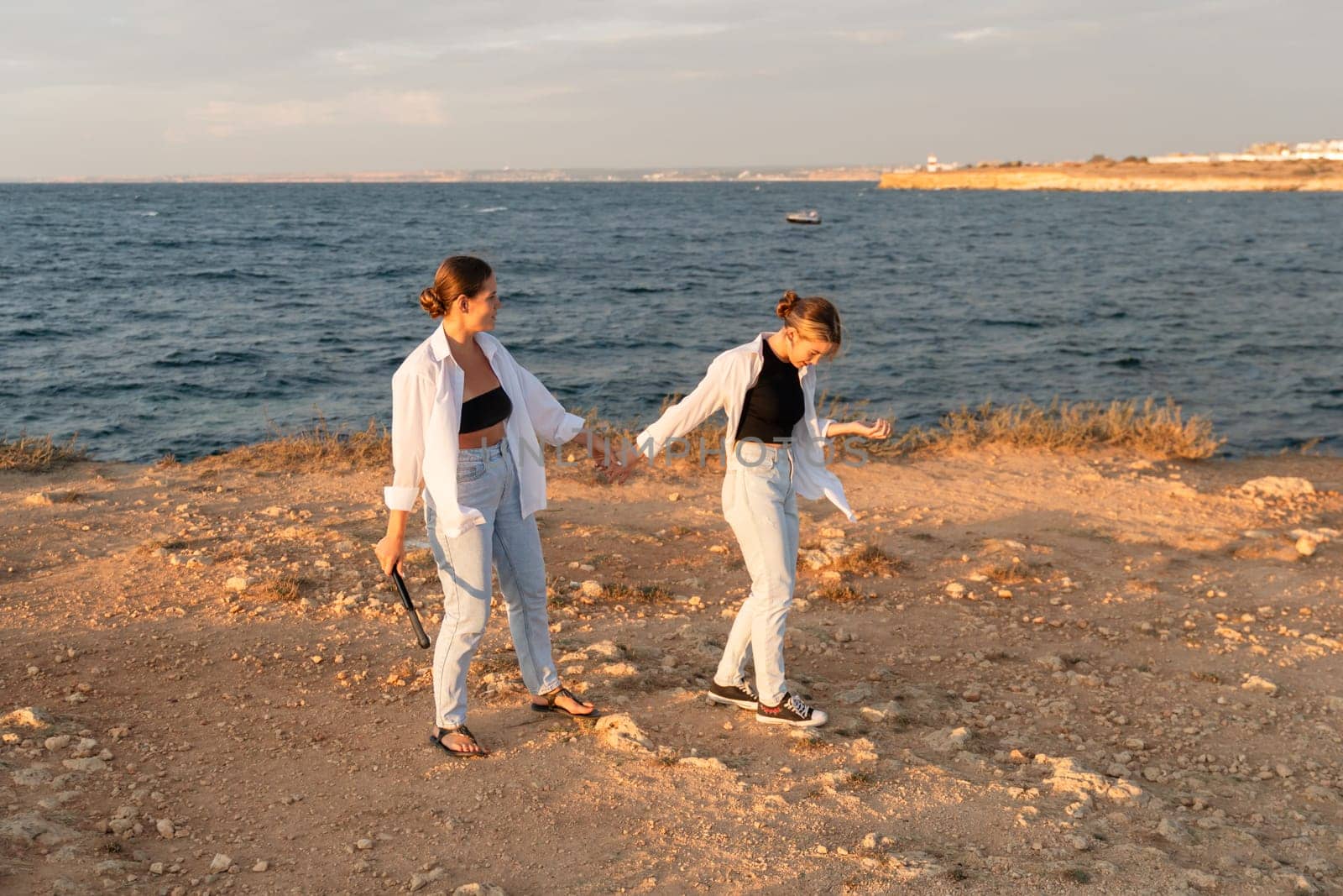 Two women in white shirts and jeans walk hand-in-hand along a rocky shoreline, the sea behind them.