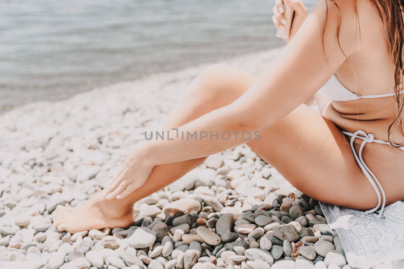 A woman is sitting on a beach with her legs crossed and a bottle of sunscreen in her hand