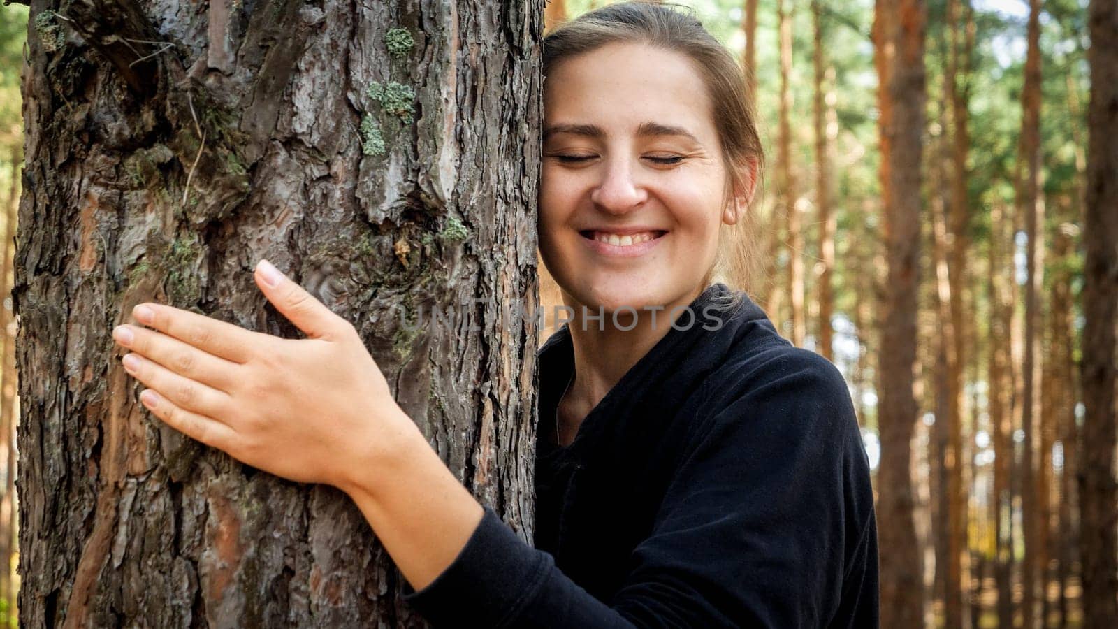 Portrait of smiling female ecology activist hugging big old tree in forest. Concept of ecology, environment protection and harmony with nature.