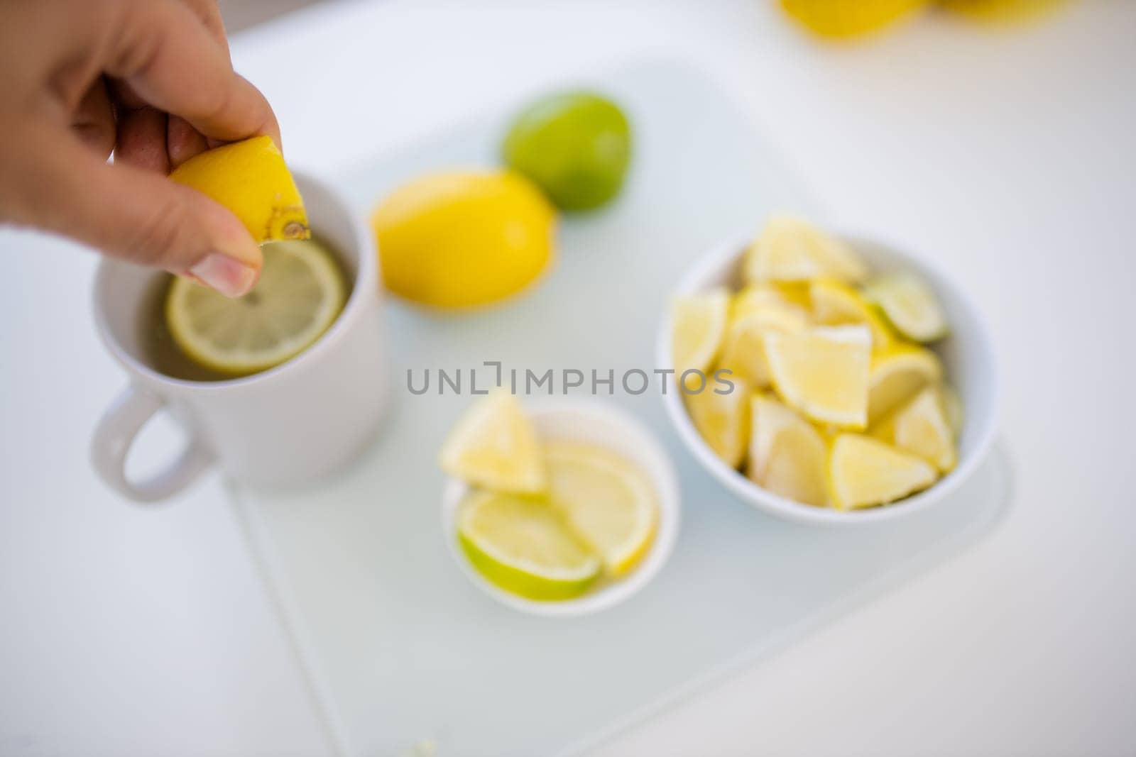 Female hand squeezing a lemon into cup of lemon tea next to lemons and limes slices. White mug and citric fruit on white table. Sweet citric beverages
