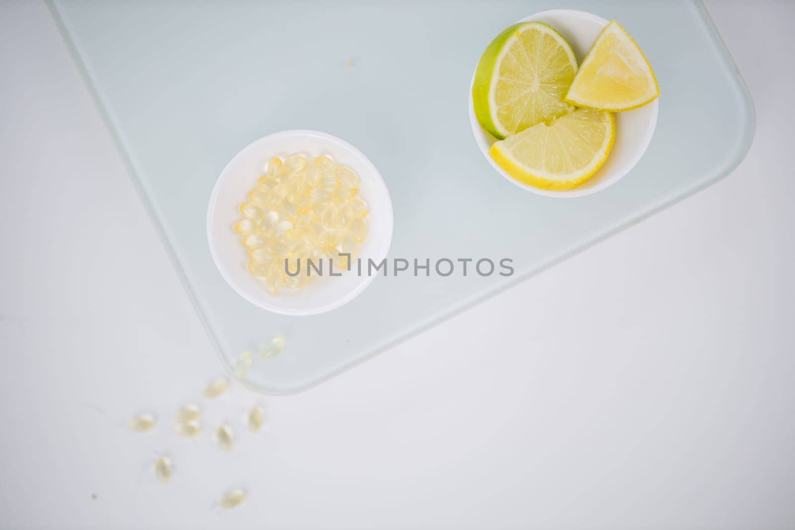 Top view of bowl of transparent vitamins next to bowl of lemon and lime slices on white table. Capsules and chopped citric fruit above transparent board. Vitamin C nutrition