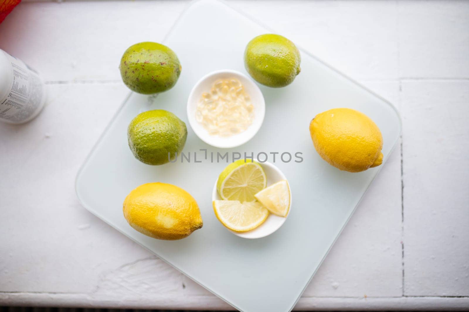 Bowl of transparent vitamins next to bowl of lemon and lime slices on white table. Capsules and citric fruit above transparent board. Vitamin C nutrition