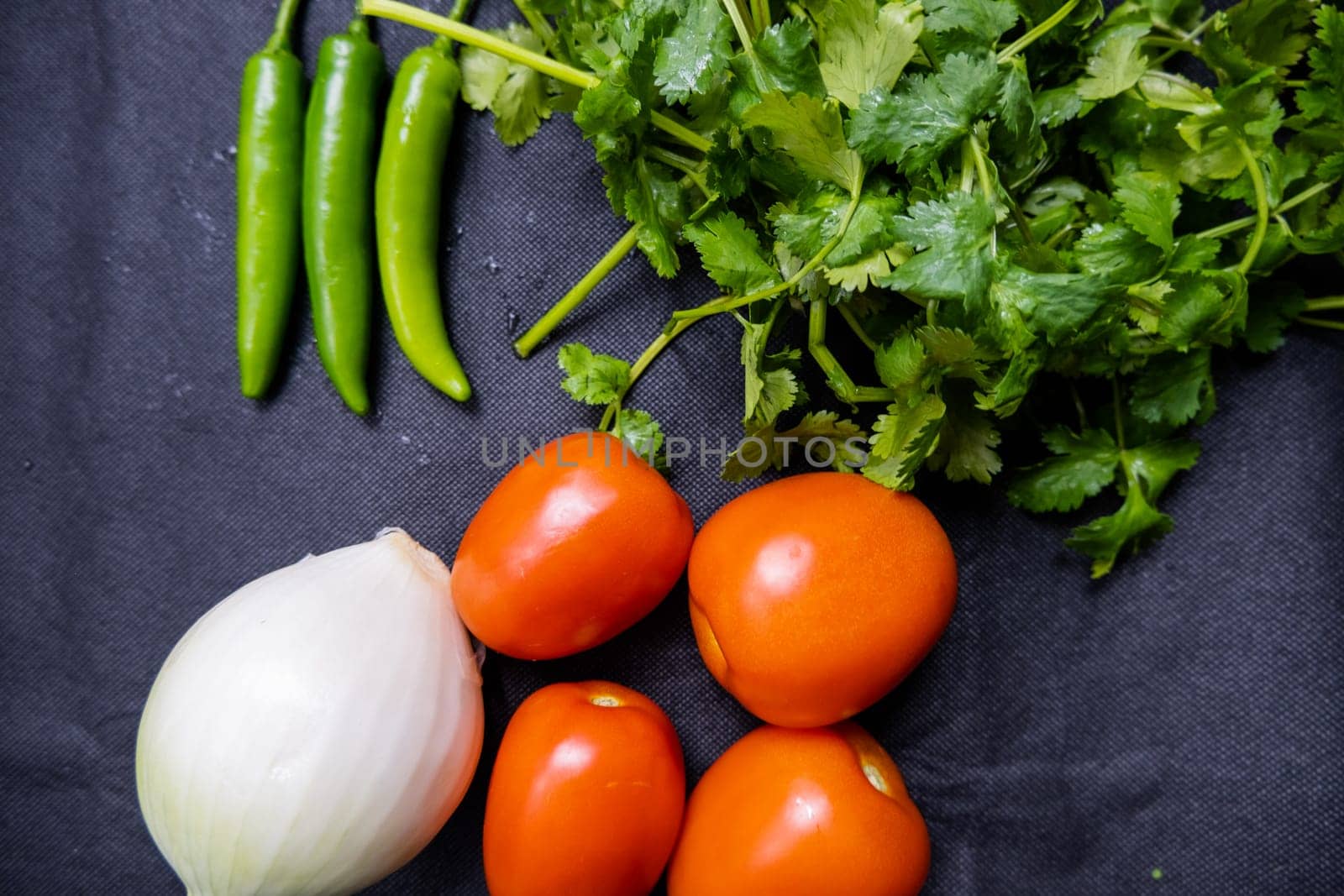 Top view of three green chili peppers, onion, four tomatoes, and coriander on table. Fresh and aromatic vegetables and herbs on dark blue surface. Healthy meal preparation