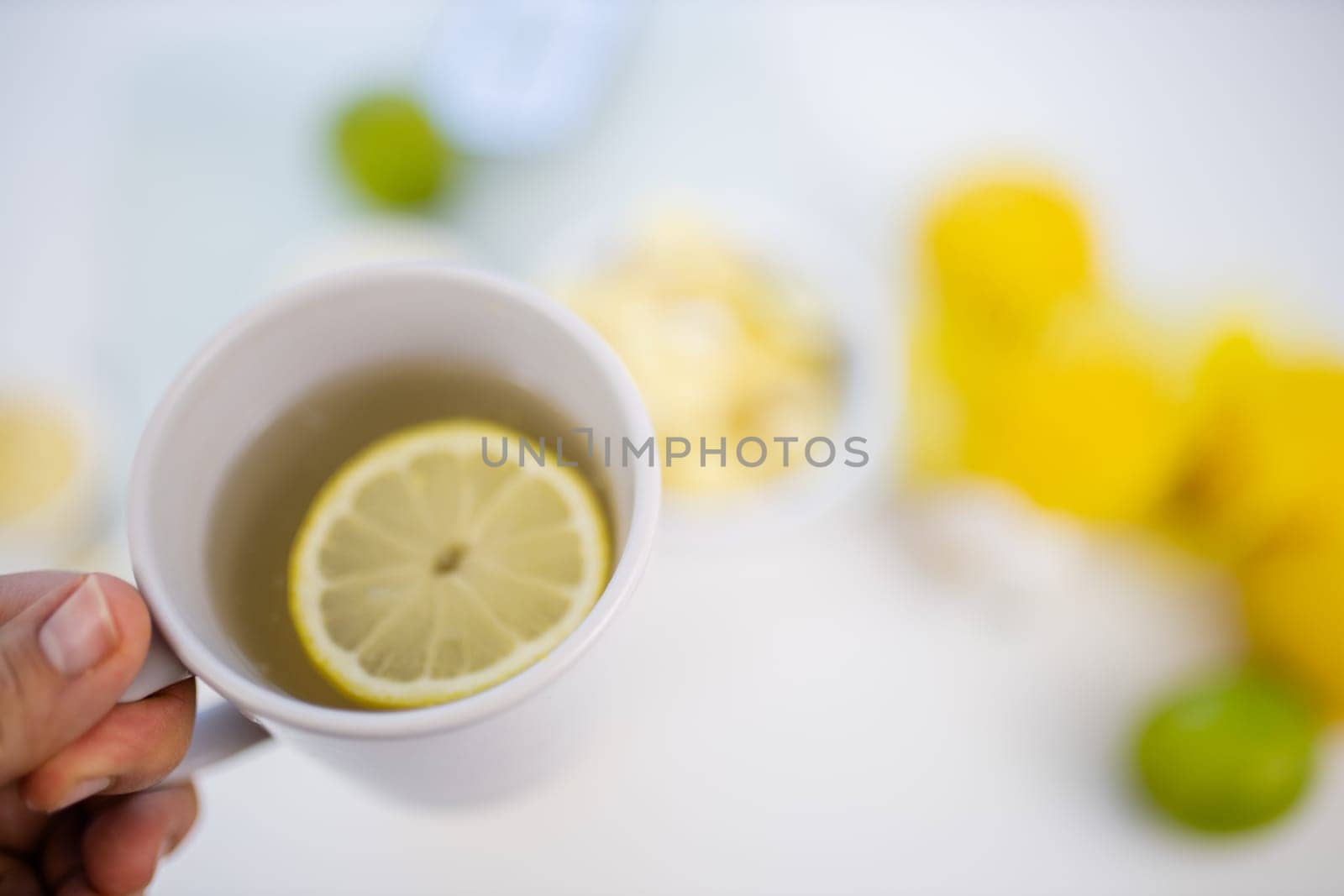 Female hand holding cup of lemon tea above blurry white table with limes and lemons. Woman holding white mug with lemon slice floating inside. Sweet citric beverages