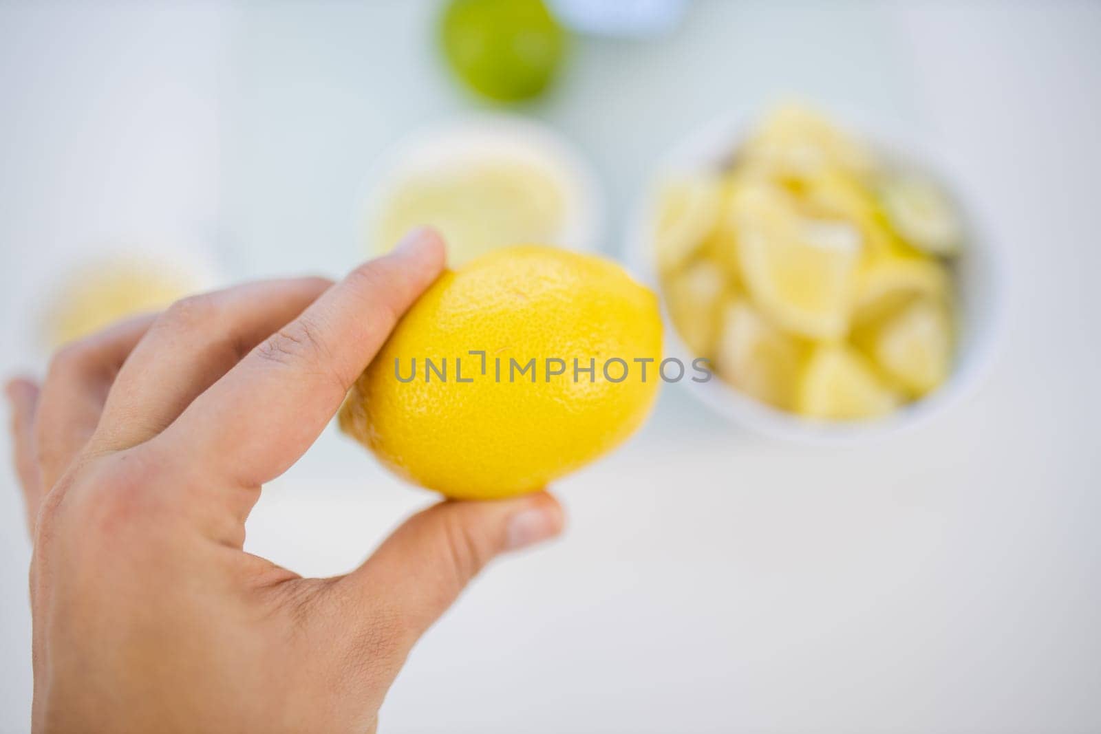 Top view of female hand holding a lemon above blurry small bowl of lime and lemon slices. Woman cutting lemons into small pieces. Citrus drink preparation