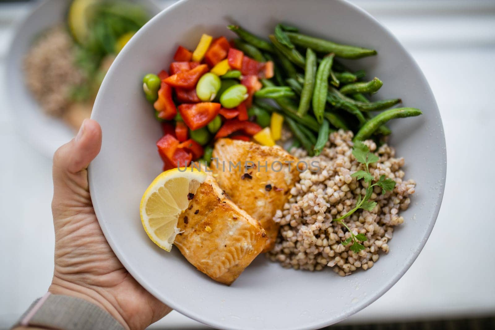 Hand holding salmon and buckwheat dish with green beans, broad beans, and tomato slices. Nutritious dish with vegetables and fish on white plate. Healthy balanced diet