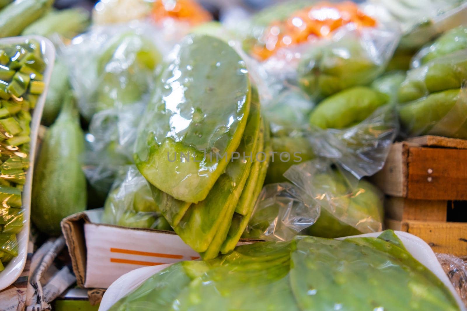 Close-up of colorful vegetable stand with sliced nopales wrapped in plastic and blurry background. Fresh-looking vegetables for sale on stall inside classic Mexican market. Natural healthy food