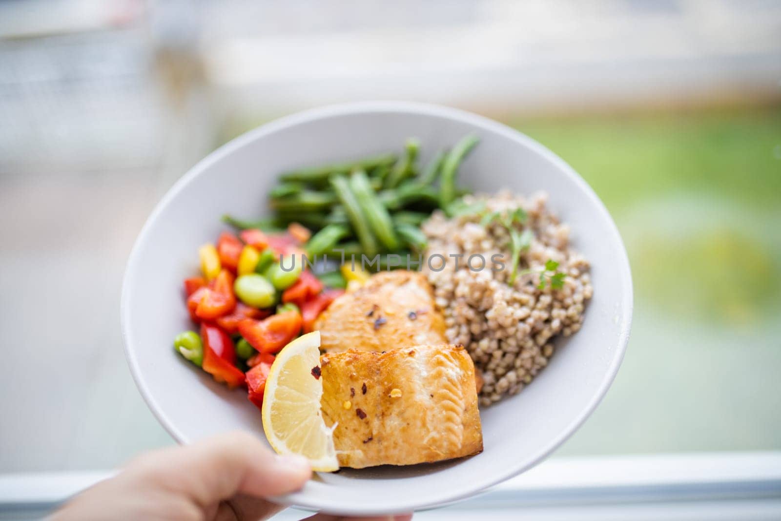 Hand holding salmon and buckwheat dish with green beans, broad beans, and tomato slices. Nutritious dish with vegetables and fish on white plate. Healthy balanced diet