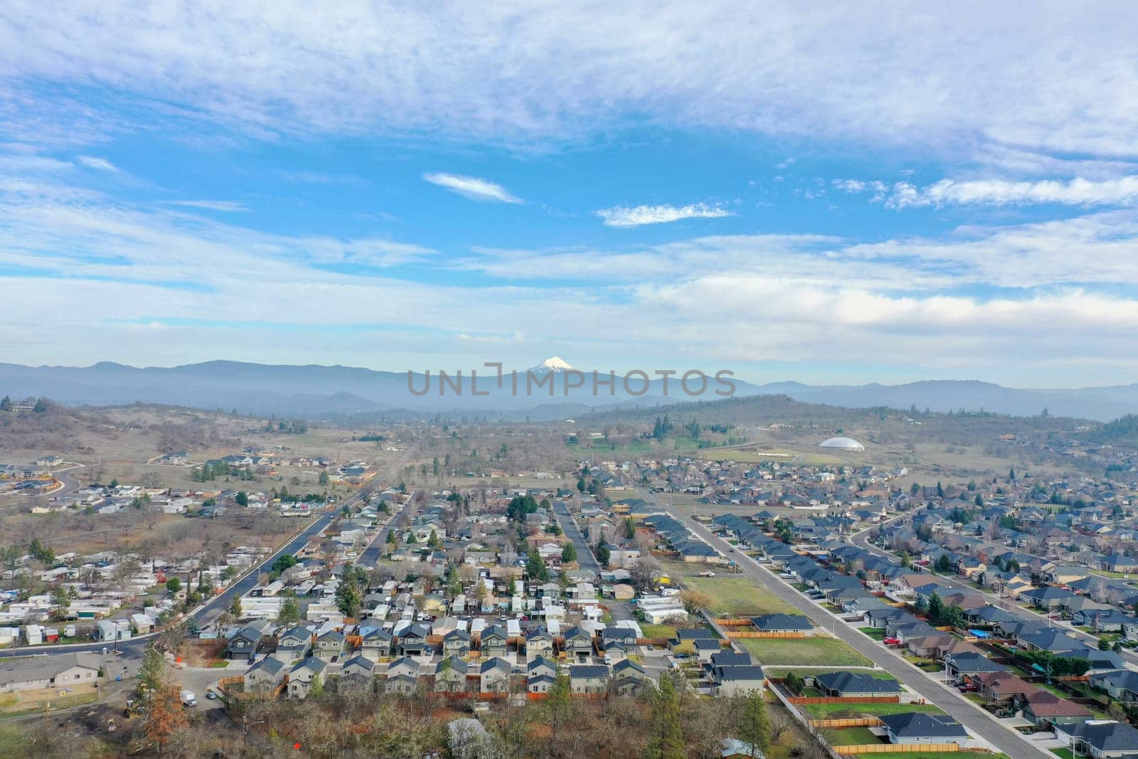 Peaceful small town under slightly cloudy sky and with mountains as background. Aerial view of slightly cloudy blue skyline above the suburbs. Rural town landscape