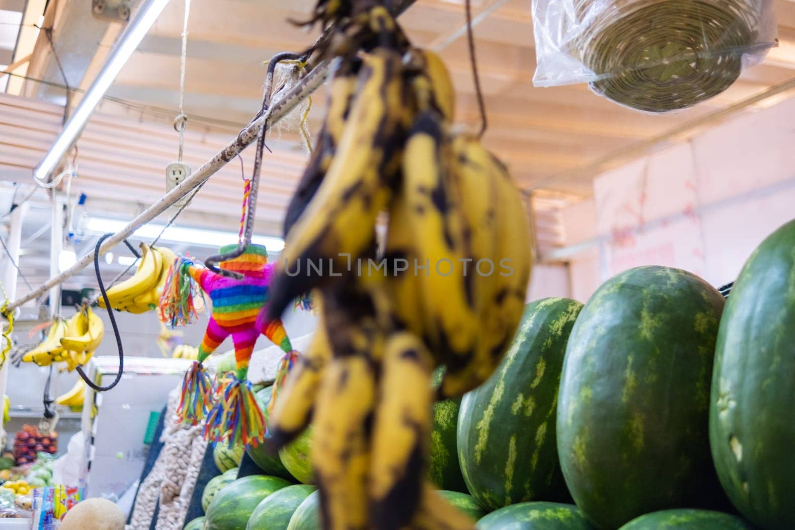 Bunch of hanging black-spotted bananas with blurry watermelons and a pinata as background. Fresh fruit with black spots for sale in classic Mexican market. Healthy food
