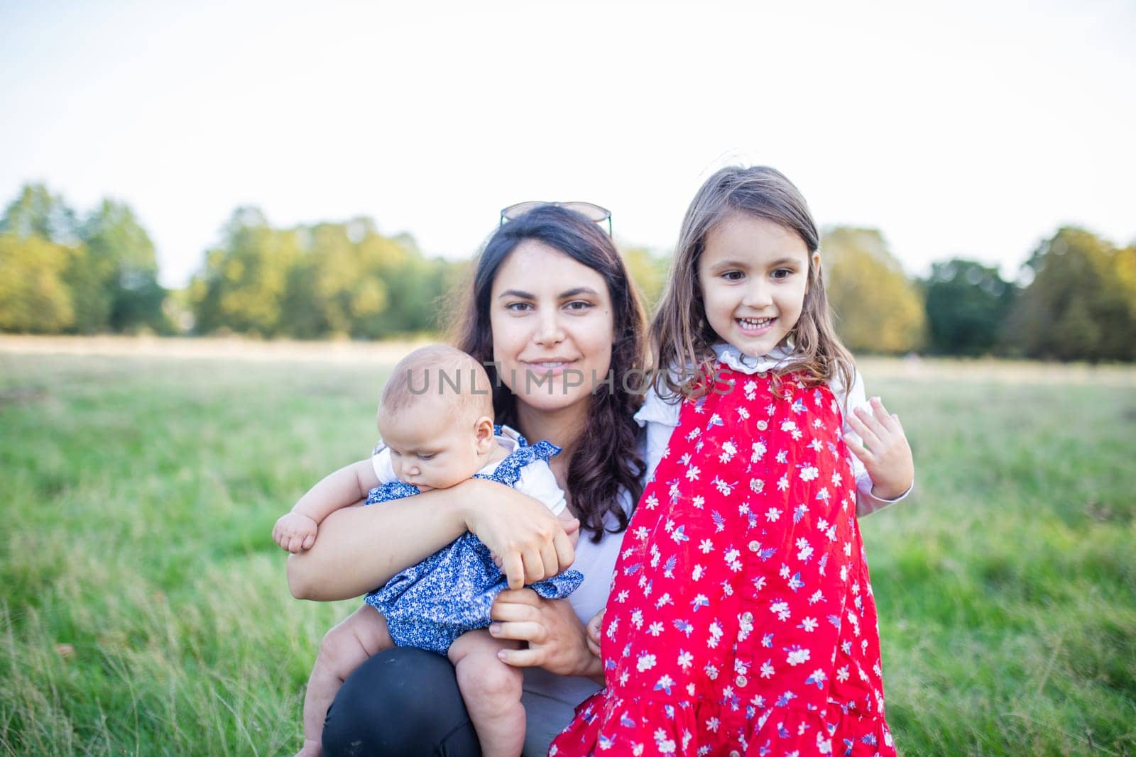Happy woman sitting on grass with cute baby and young girl with trees as background. Mother hugging adorable daughters in park. Young family having fun outdoors