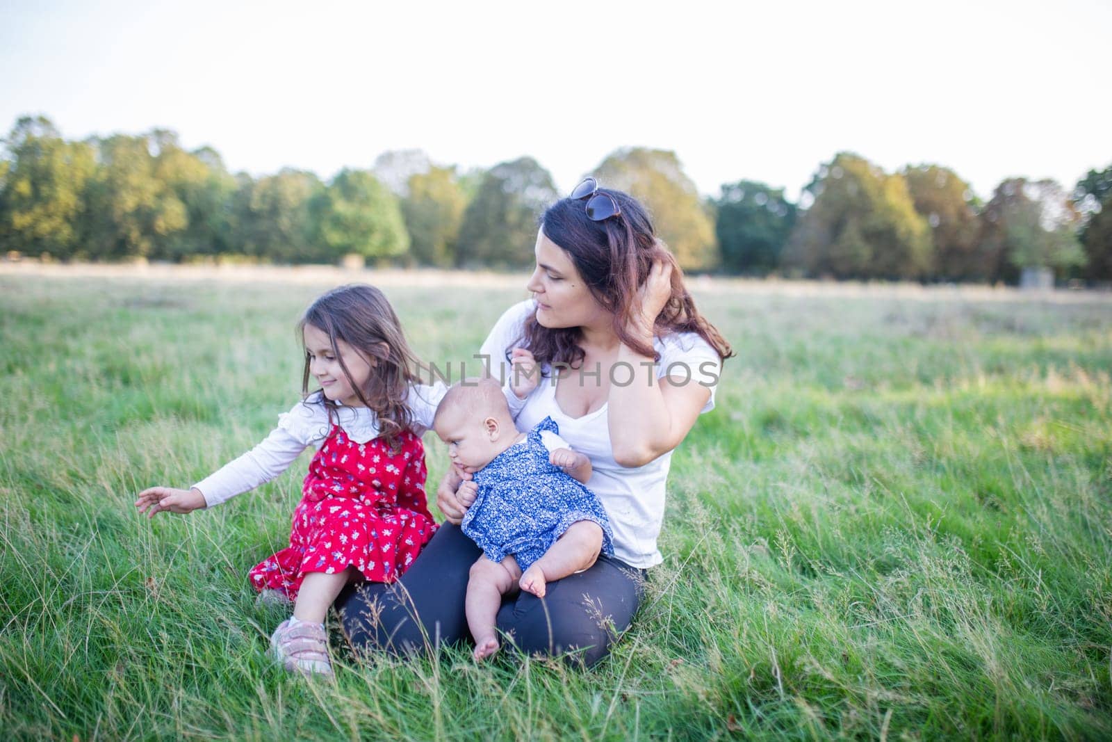 Happy woman, baby, and little girl sitting on grass with trees as background. Mother playing with adorable young daughters in park. Young family having fun outdoors