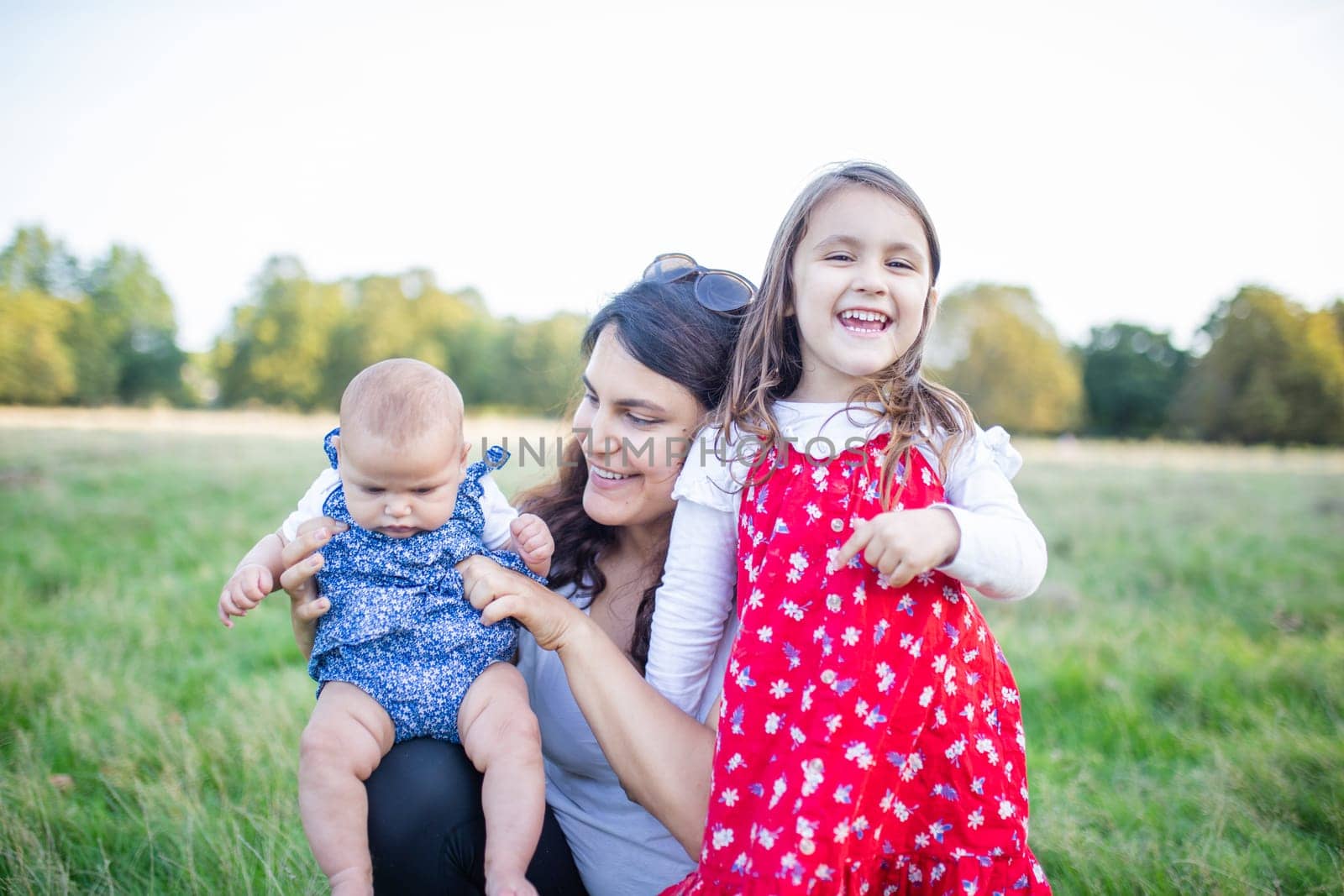 Happy woman, baby, and little girl sitting on grass with trees as background. Smiling mother holding adorable daughters in park. Young family having fun outdoors