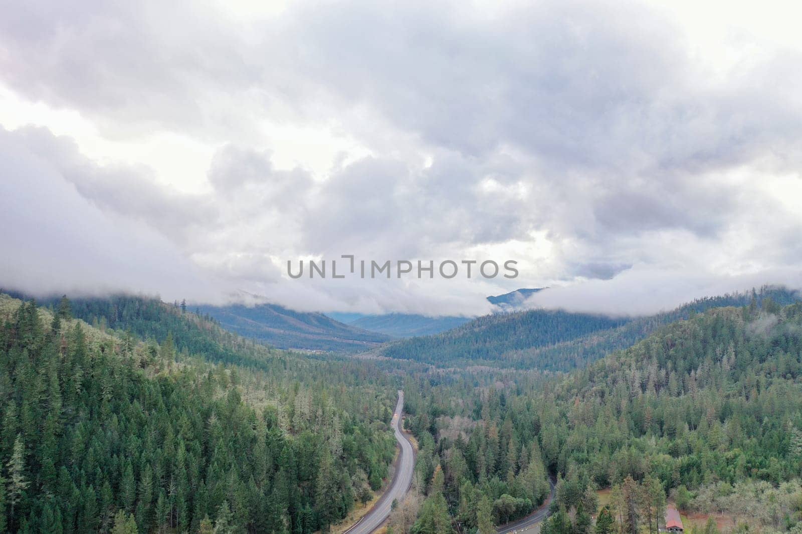Beautiful and peaceful forest under cloudy sky and with mountains as background. Aerial view of road in the middle of the woods. Wooded and rocky landscapes