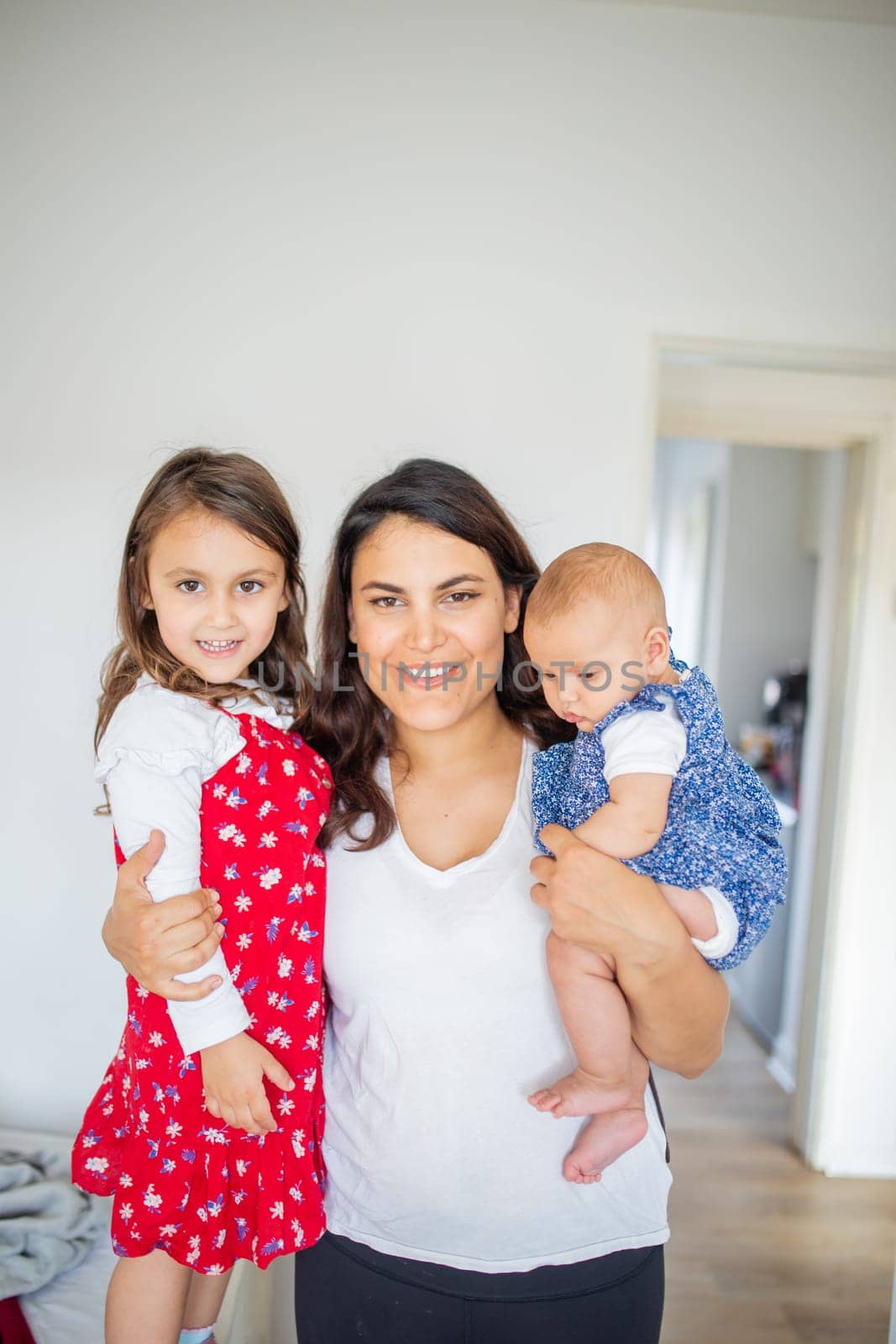 Happy woman inside white bedroom lovingly hugging her adorable young daughters. Smiling mother tightly holding cute little girl and baby. Young family at home