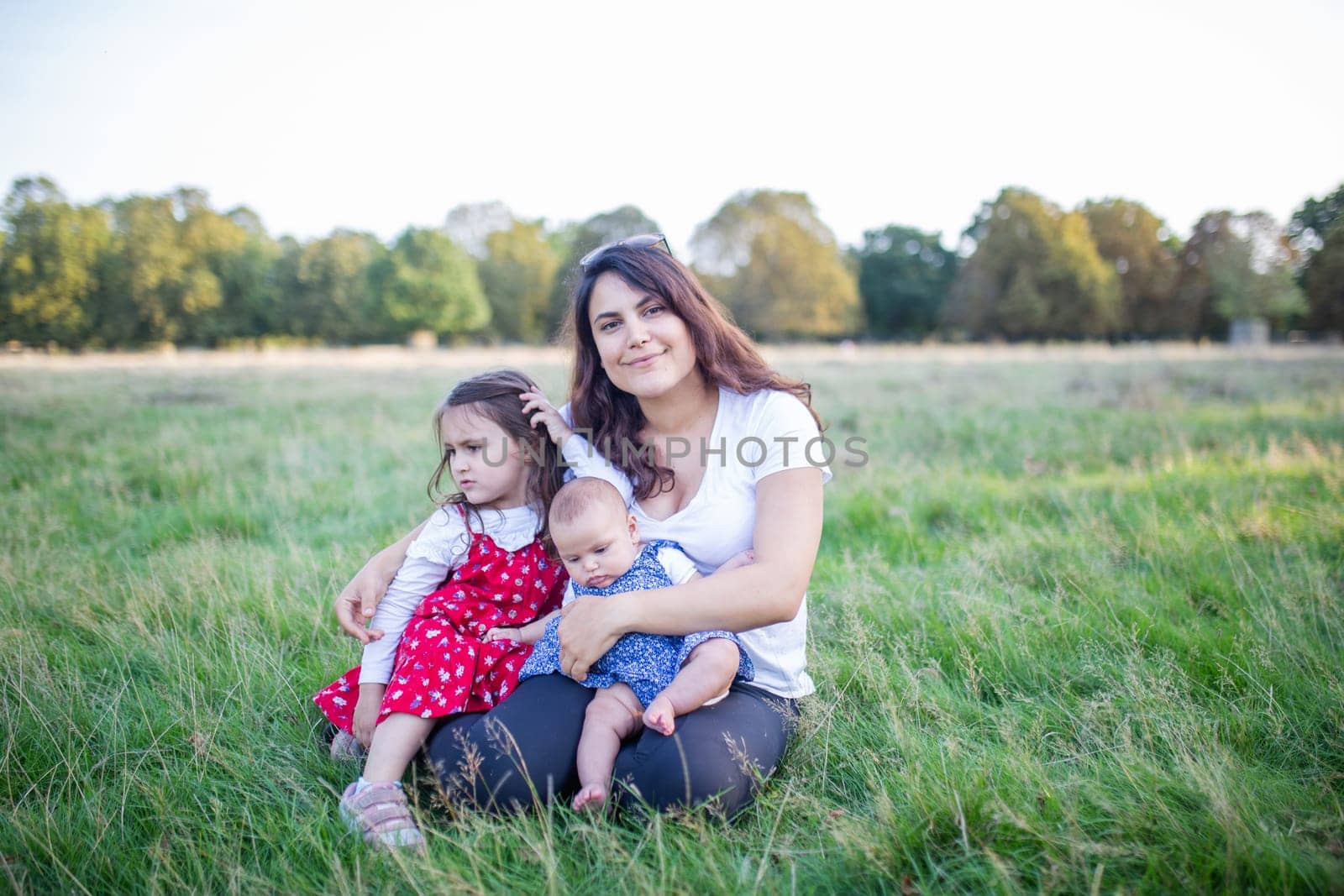 Happy woman, baby, and little girl sitting on grass with trees as background. Smiling mother hugging adorable daughters in park. Young family having fun outdoors