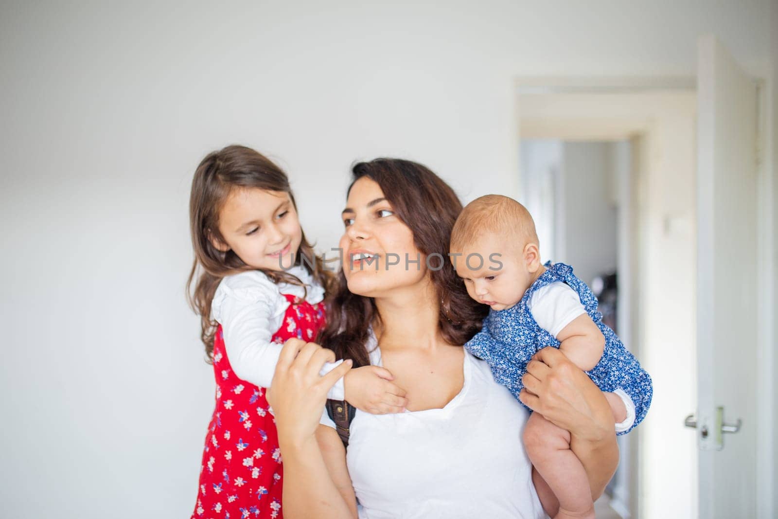 Happy woman inside white bedroom lovingly hugging her adorable young daughters. Smiling mother hugging cute little girl and baby. Young family at home