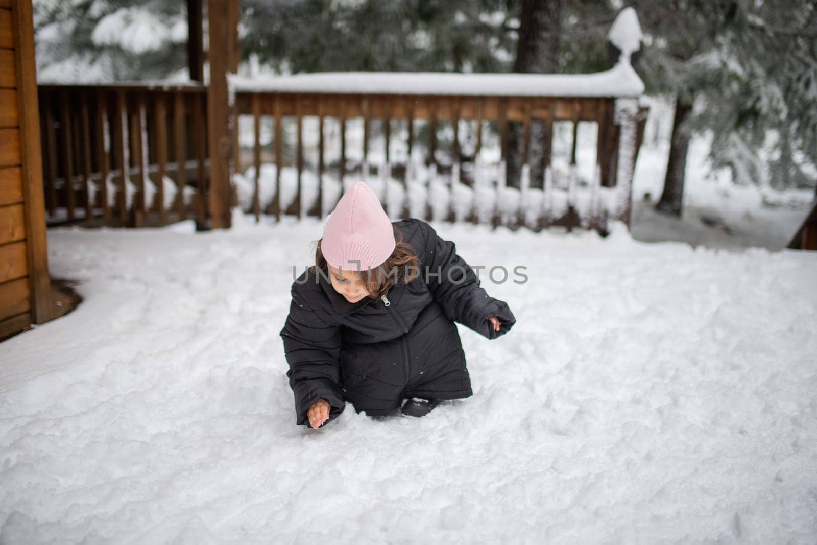 Happy little girl wearing oversized jacket kneeling and playing on the snow. Young child having fun on snowy day. Adventurous winter holiday